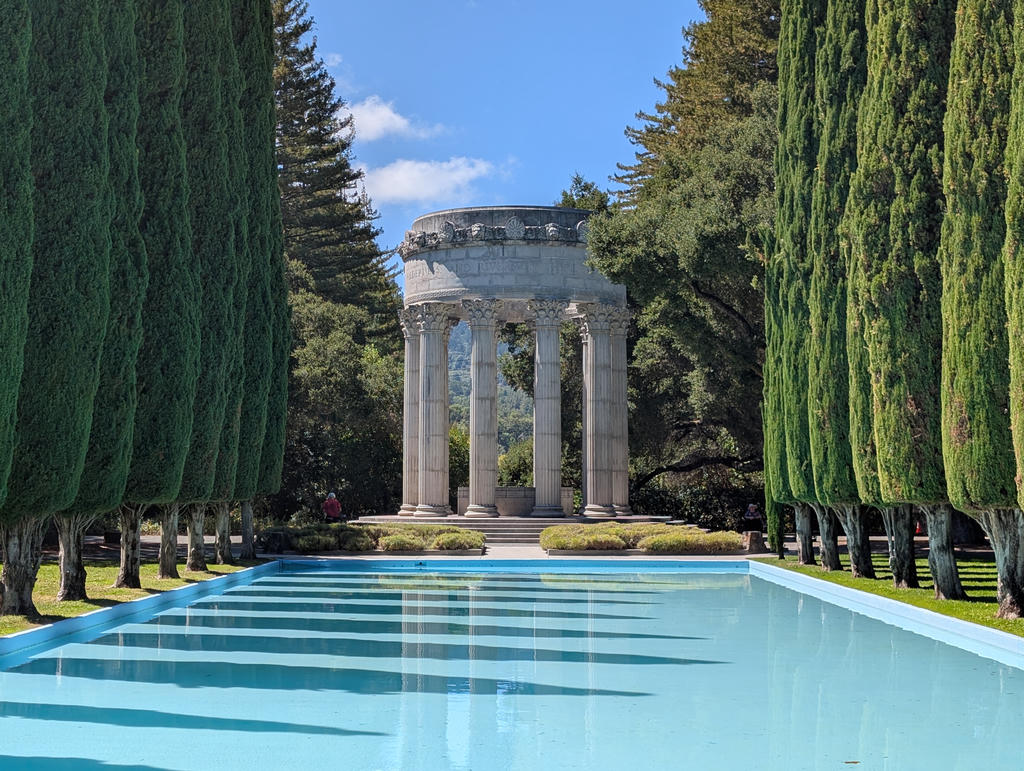 A Roman-esque round temple, with a shallow pool in the foreground. Two rows of skinny trees are on either side of the pool.